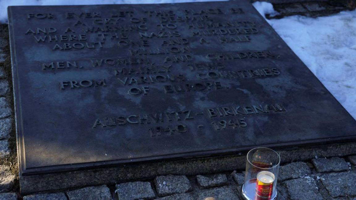 A candle in a clear holder is burning in front of a plaque at Auschwitz-Birkenau, which is surrounded by snow.