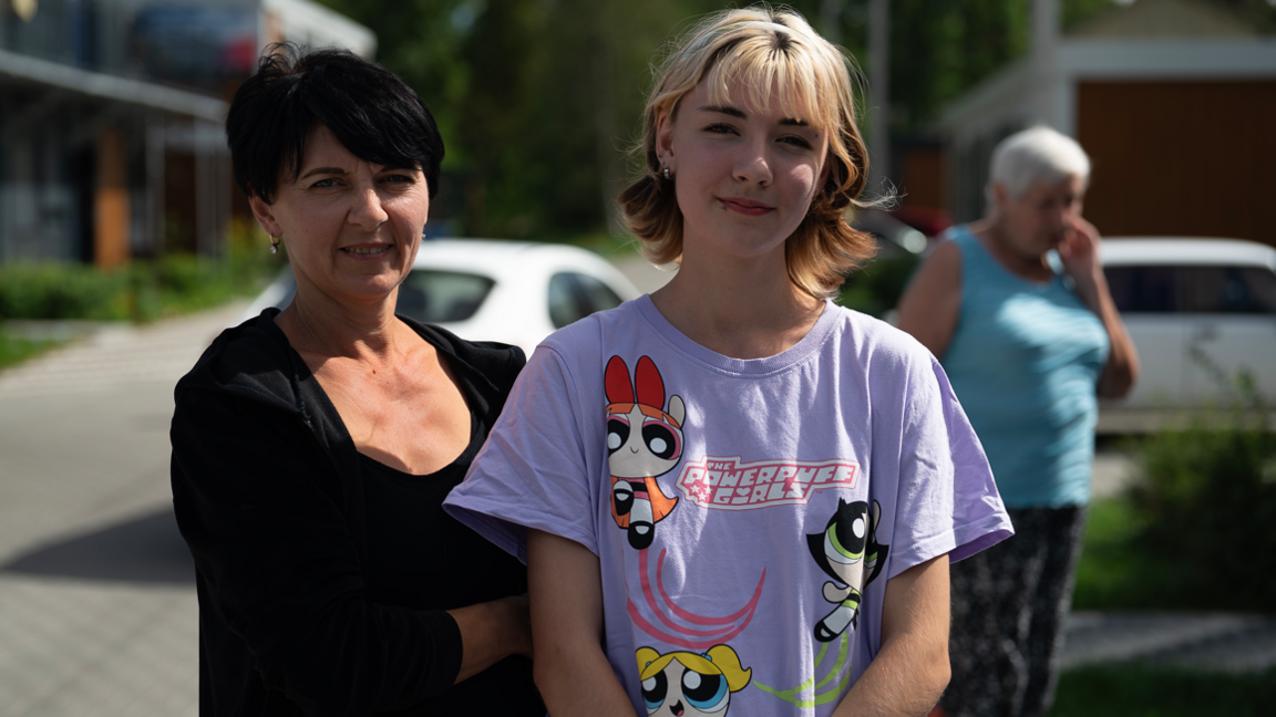 Valeria (right) stands in a lilac Powerpuff Girls t-shirt with her mother in their refugee village