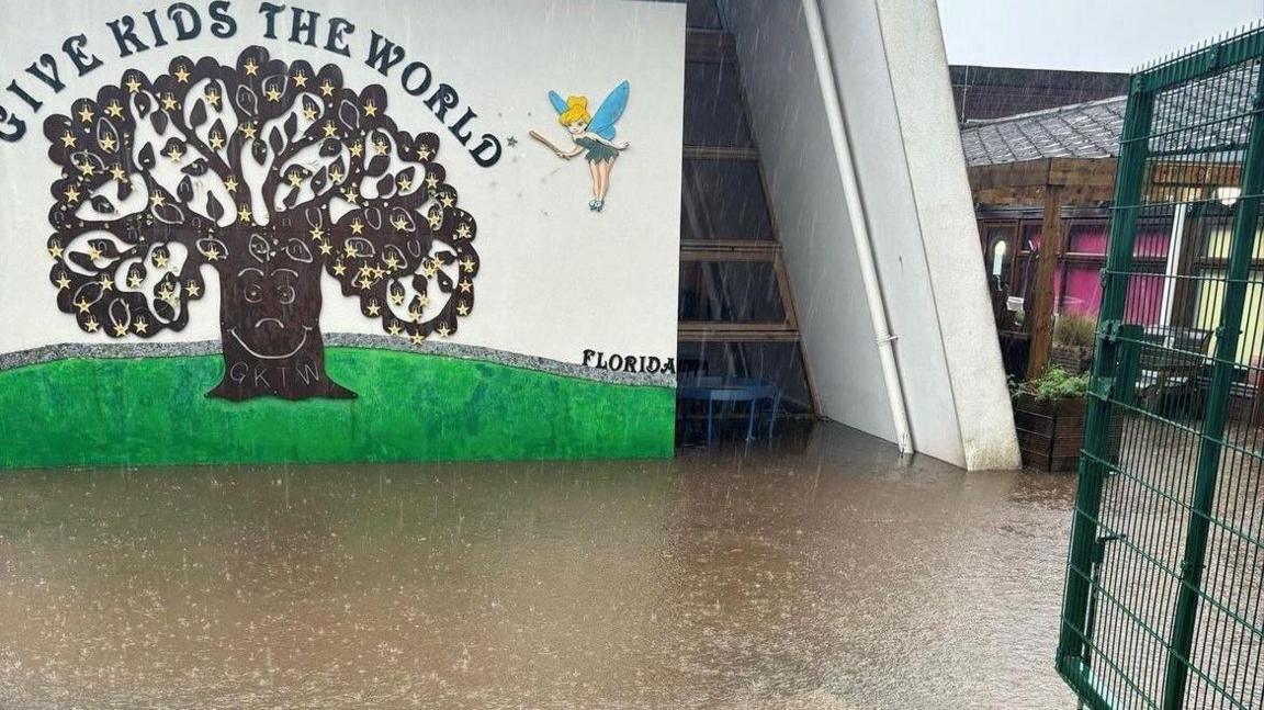 Heavy rain outside a school entrance with the words give kids the world and a picture of a tree on a white wall