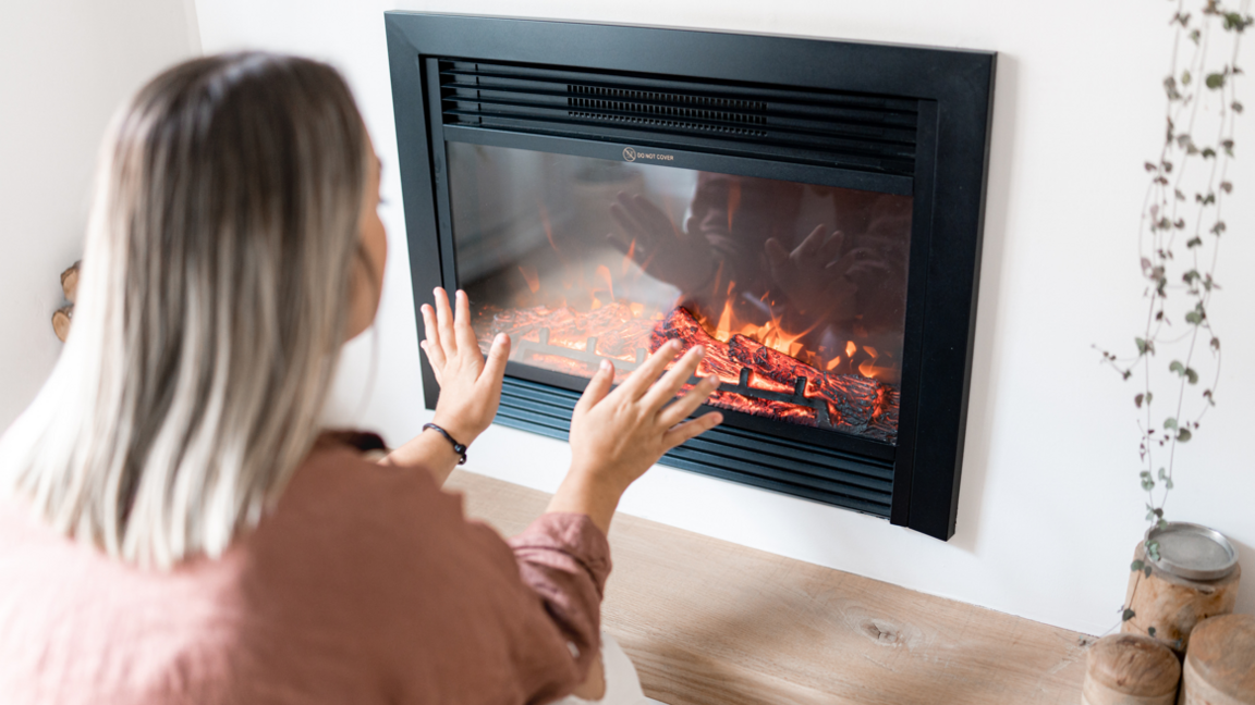 Woman with blonde hair in pink-ish top warms her hands on a gas fire 