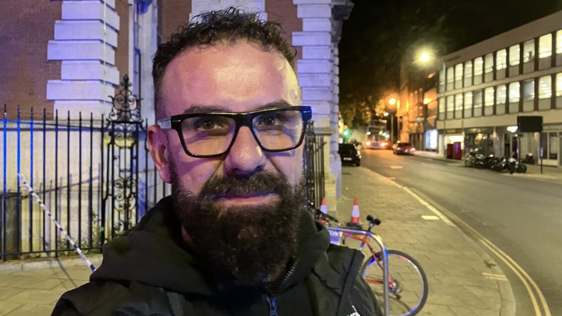 Mr Ademaj looks towards the camera. He has short black hair, a long black beard and is wearing black glasses and coat. Behind him to the right is a road with buildings on either side.