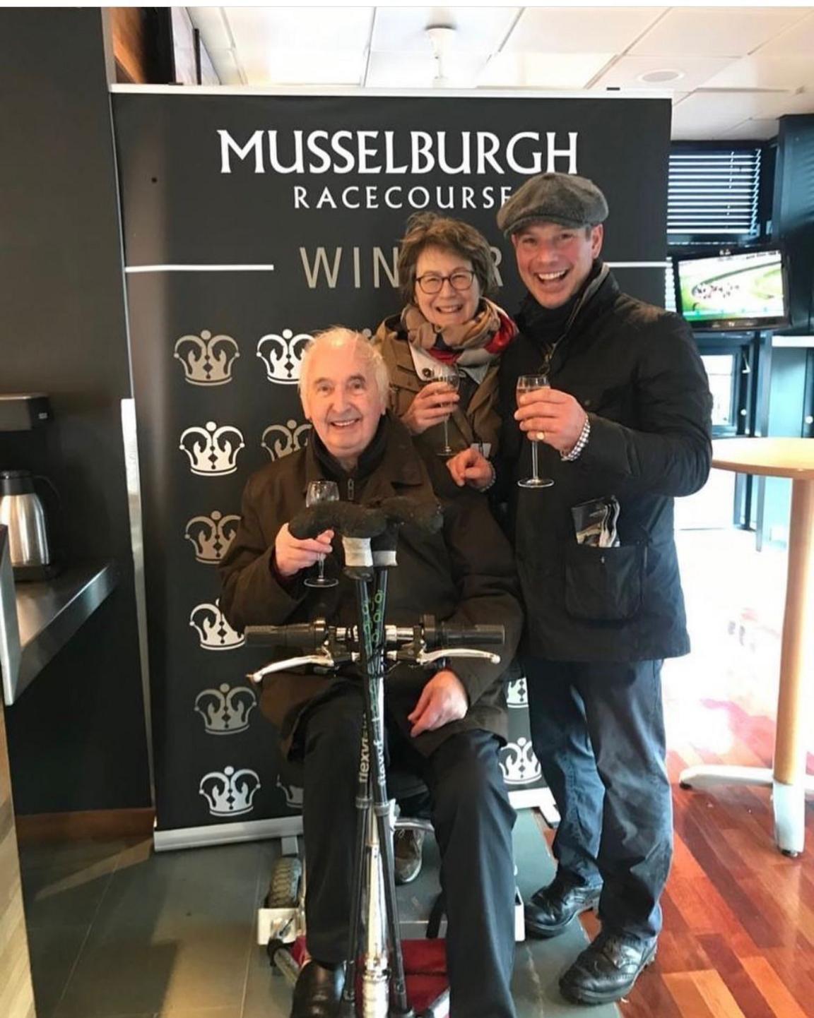 Three people, one sitting down and two standing up raise glasses of champagne to celebrate a winning horse at Musselburgh Racecourse
