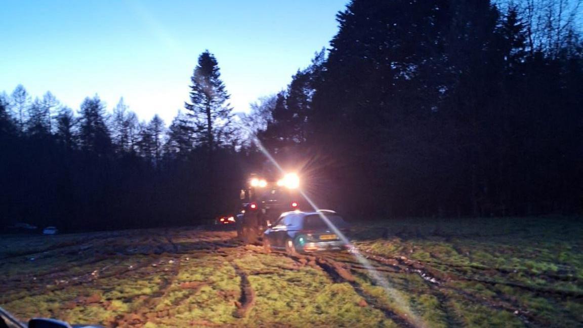 A blurry photo of a tractor towing a car across a muddy field. It's getting dark and the tractor's headlights are on.