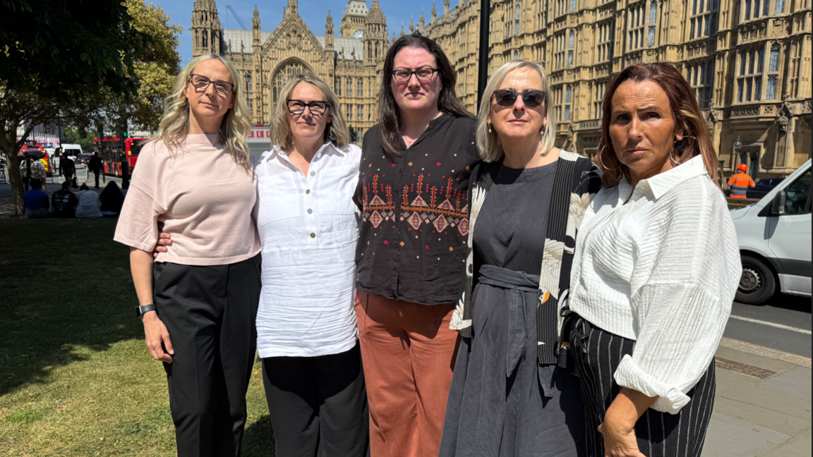 Five women stood holding each other by the waist and facing the camera. They are stood outside Parliament.