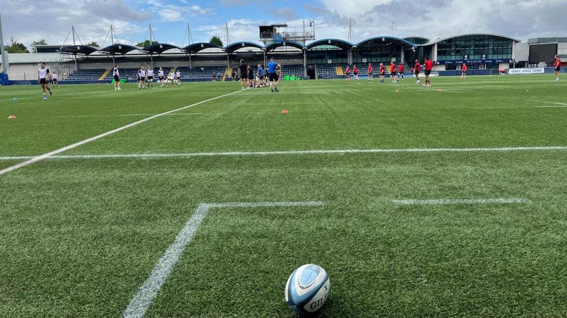 A rugby ball is at the bottom of the photo. There is a team in white on the left on the pitch and players in red are on the right. A stand is in the distance