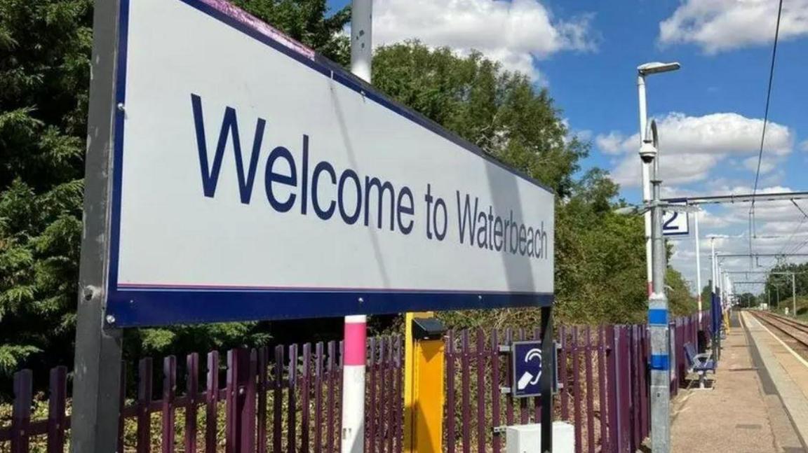 A sign saying 'Welcome to Waterbeach' above purple railings and a train platform and track to the right.
