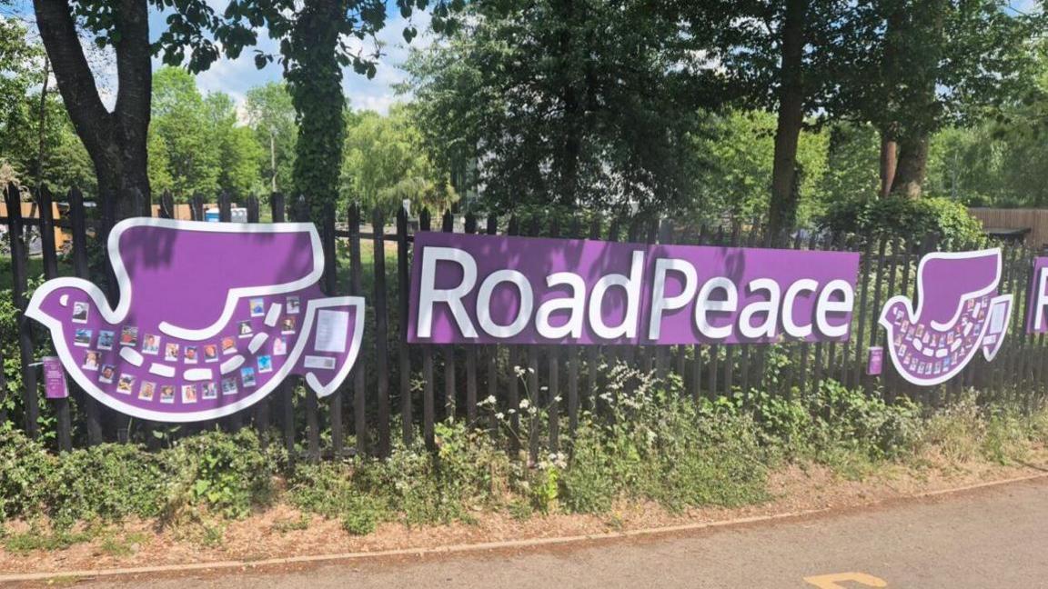 A large purple sign with Road Peace in white lettering hangs on a metal fence with trees behind. There are two purple doves, one on either side, with photos attached to them.