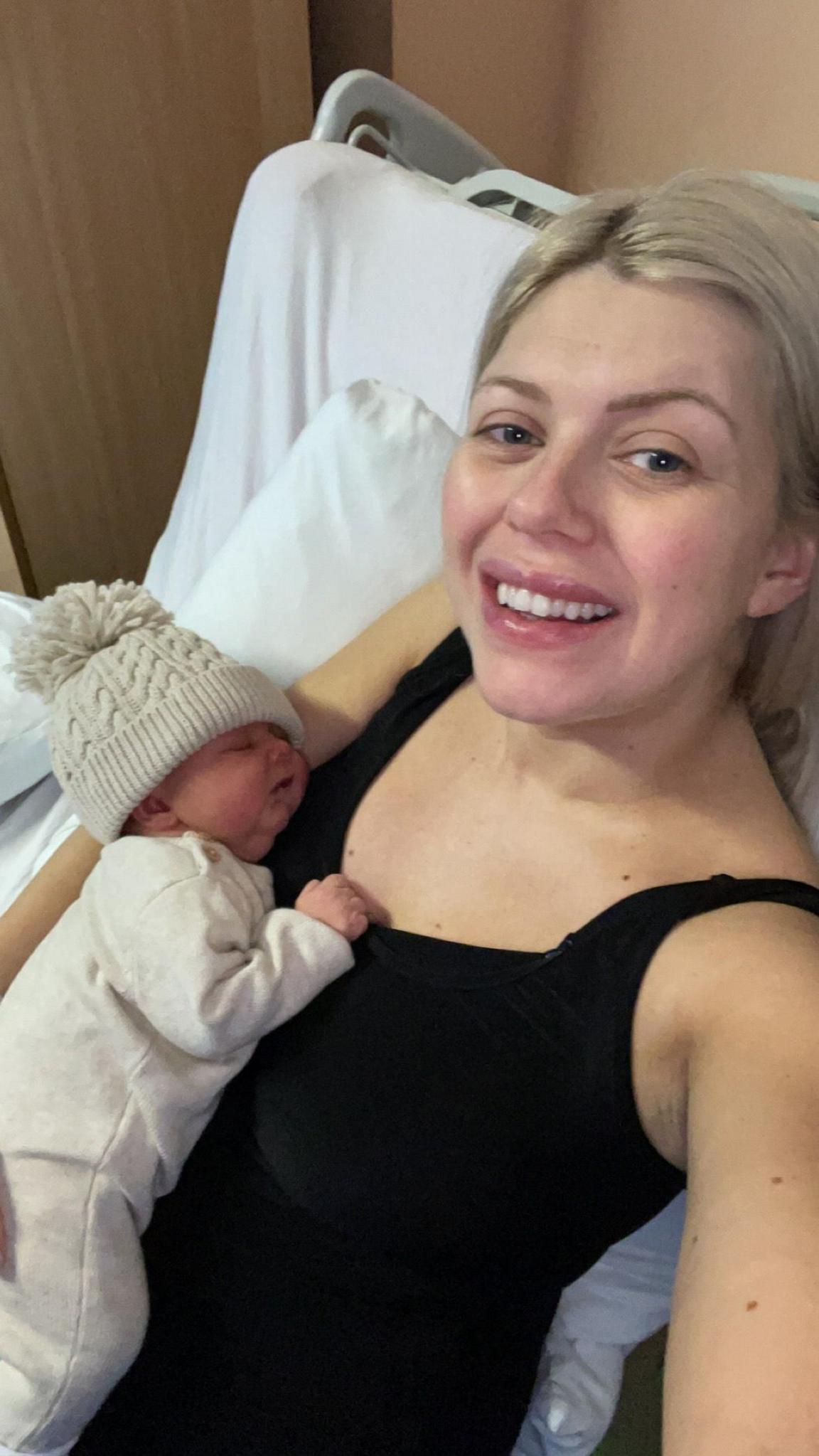 Julie Scott McLean in a black vest top with blonde hair taking a selfie of herself and a newborn baby in white top and cream woolly hat.