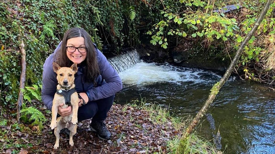 Melanie Warburton and her dog Winston in a wooded area by a stream. Melanie is wearing a purple coat and blue jeans and is crouched looking at the camera. She has long dark hair and is wearing black-rimmed glasses.