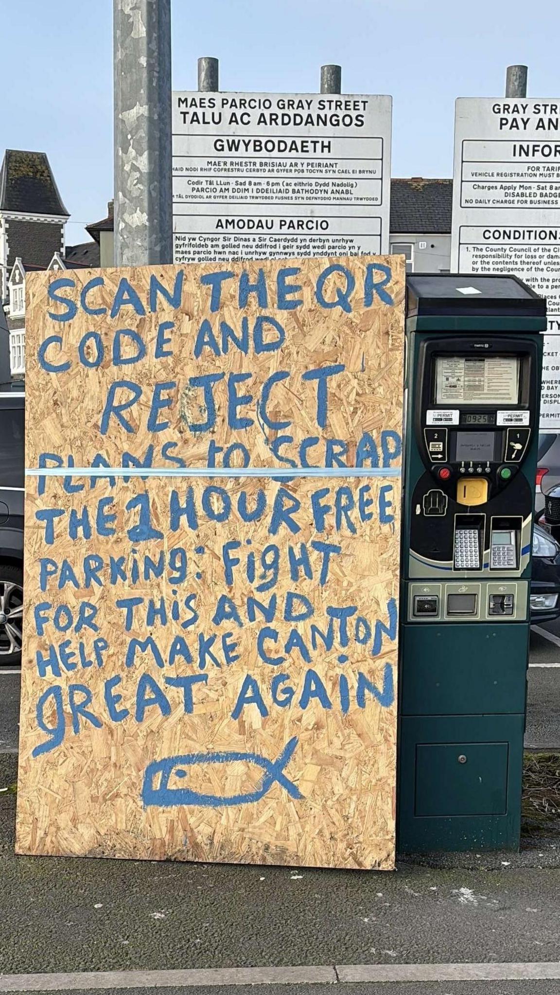 A large sign propped up against a parking meter in a Canton car park that reads "scan the QR code and reject plans to scrap the 1 hour free parking: fight for this and help make Canton great again'. 