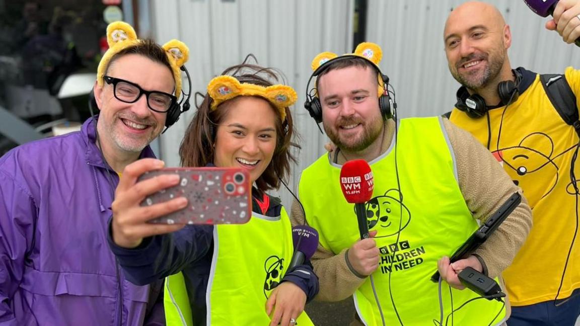 Four people - three men and a woman - are wearing yellow Pudsey Bear ears on their heads and yellow high-vis jackets are posing for a selfie. Some of them are holding microphones