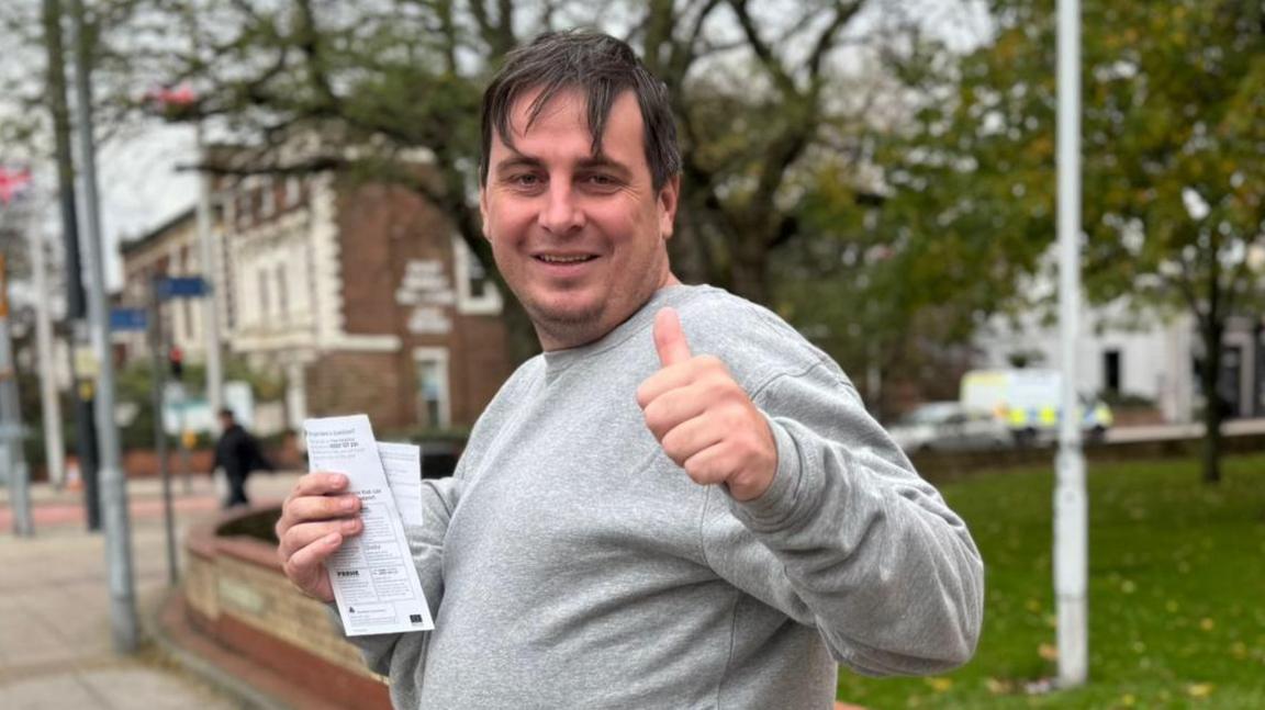 Daniel Jarvis, 37, who is wearing a light grey sweatshirt and jogging bottoms, smiles and gives a thumbs up to the camera.