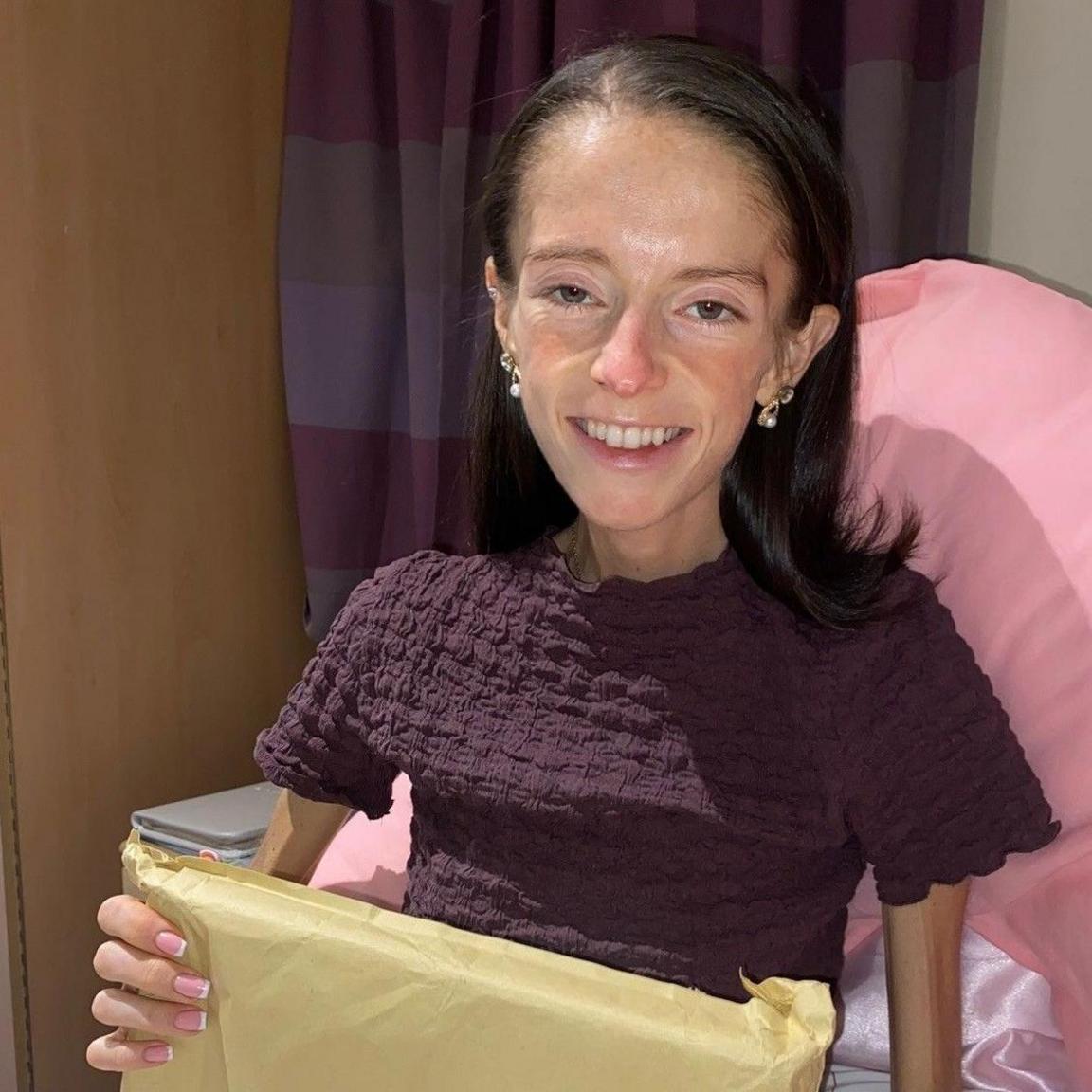 Hayley Morrison lies on top of a bed with pink bedding. She wears a brown top and has dark shoulder length hair. She holds a parcel on her lap.