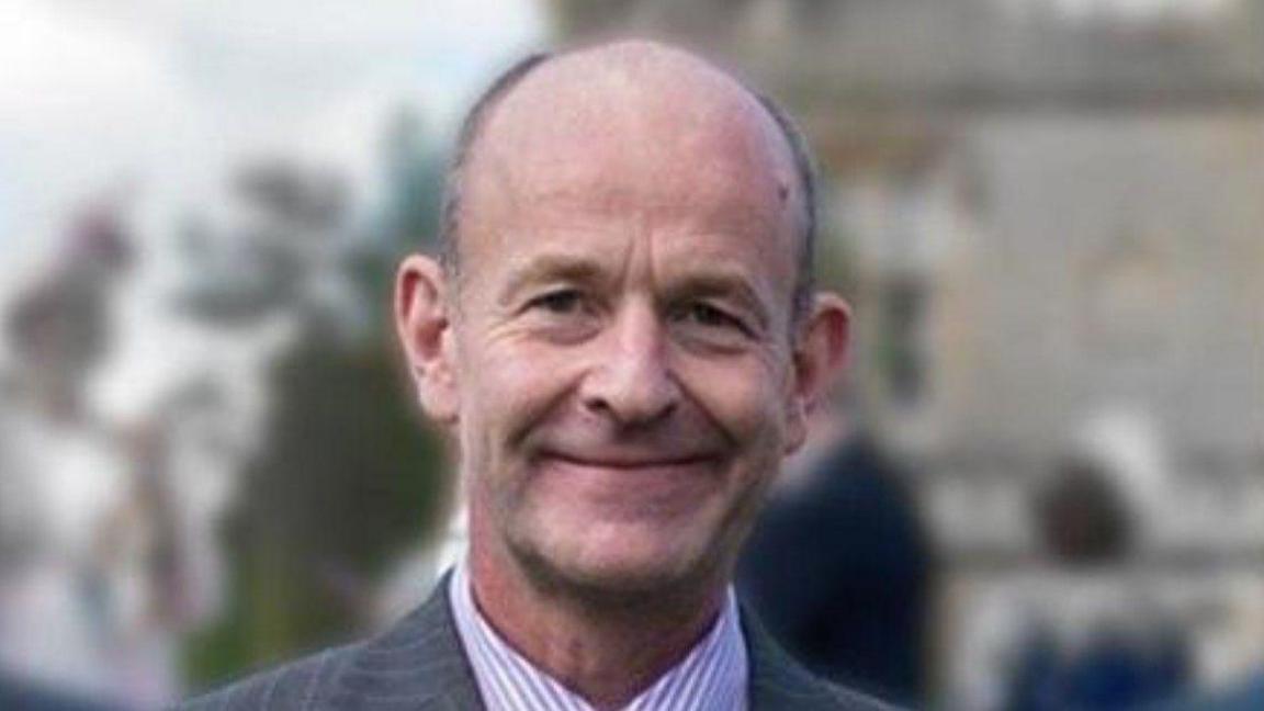 A man wearing a jacket and striped shirt is smiling at the camera. The background is blurred.