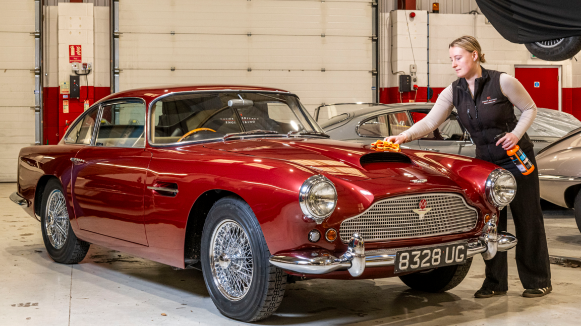 The front of a 1961 Aston Martin DB4 is being waxed by an employee at Haynes Motor Museum in a large warehouse. The car is a brilliantly-shiny red colour and has the classic, beautiful streamlined shape of an Aston Martin. 