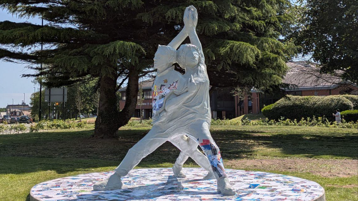 The grey sculpture, which is two people intertwined with their hands joined above their heads. Colourful patterns can be seen in sections through the sculpture. It is on a patch of grass with a road behind it.