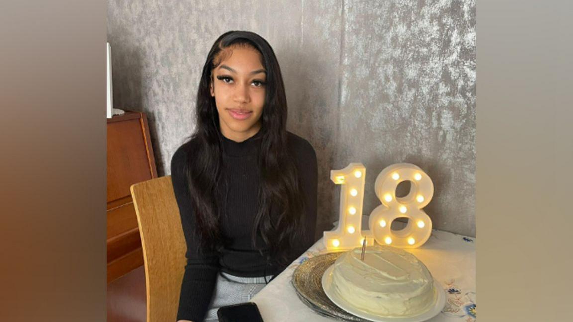 Eliza on her 18th birthday, beside a cake and a light up sign that says 18