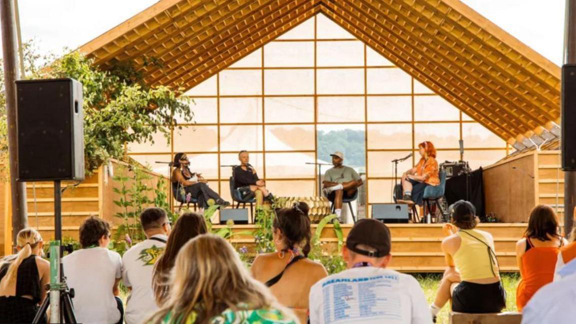 The Information stage at Forwards Festival. It is a large triangular wooden structure with wide steps leading up to the stage and thin panels at the back. There is a small crowd of people watching as four speakers on stage discuss issues