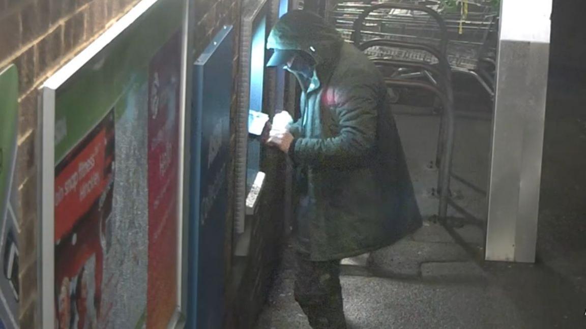 A person wearing a hooded coat and cap is using an outdoor ATM at night. The scene is dimly lit, with bright light coming from the ATM screen. There are posters on the brick wall to the left and a stack of shopping carts in the background.