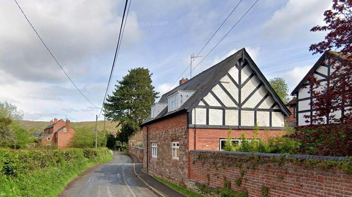 A cottage stands near a road with a walled garden. The walls are partly brick and partly black and white. Across the road, there is a grassy verge and hedge. Further cottages are around the property. There is open countryside further away.