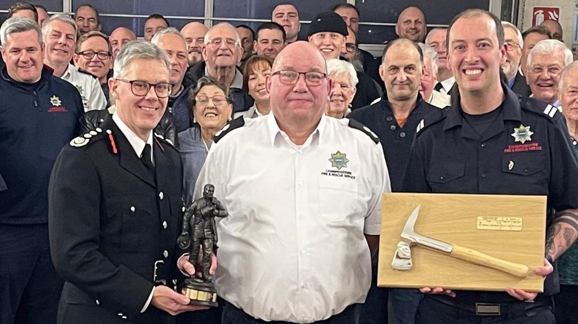 Russell Gooderham standing in the centre of a fire station surrounded by fire crews. He wears a white shirt with black shoulder epaulettes - part of his uniform. He is being presented with a momento of a firefighter's axe on a wooden board, and also a figurine of a fireman.