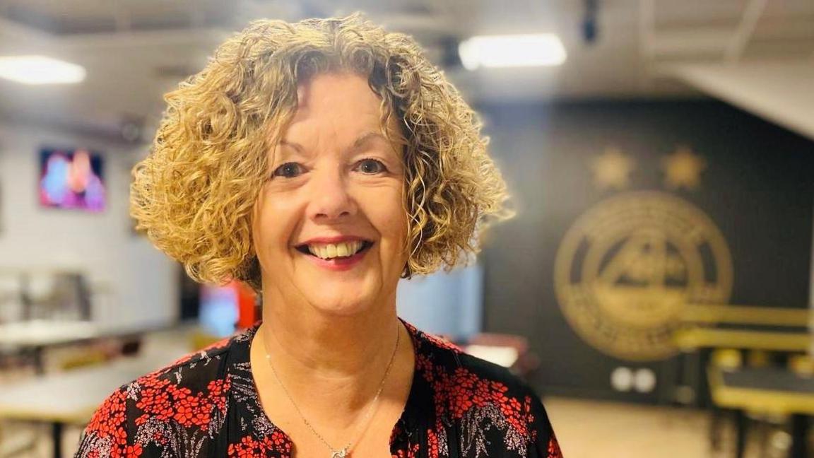 Woman with middle-parted blonde curly hair, smiling at camera, wearing a black and red top and a necklace, inside a room, with a black and gold Aberdeen Football Club blurred on a wall in the background.