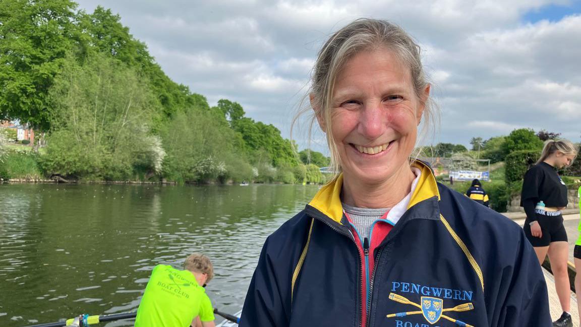 A woman with grey hair and a blue zip-up top with the word Pengwern on it stood in front of a river with a few people getting into boats behind her