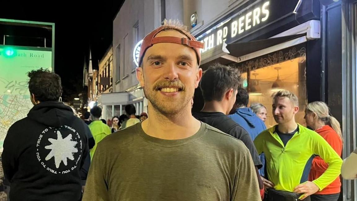 A man dressed in a green t-shirt, which a red cap on backwards, is stood outside a bar. Behind him is a group of people wearing running gear.