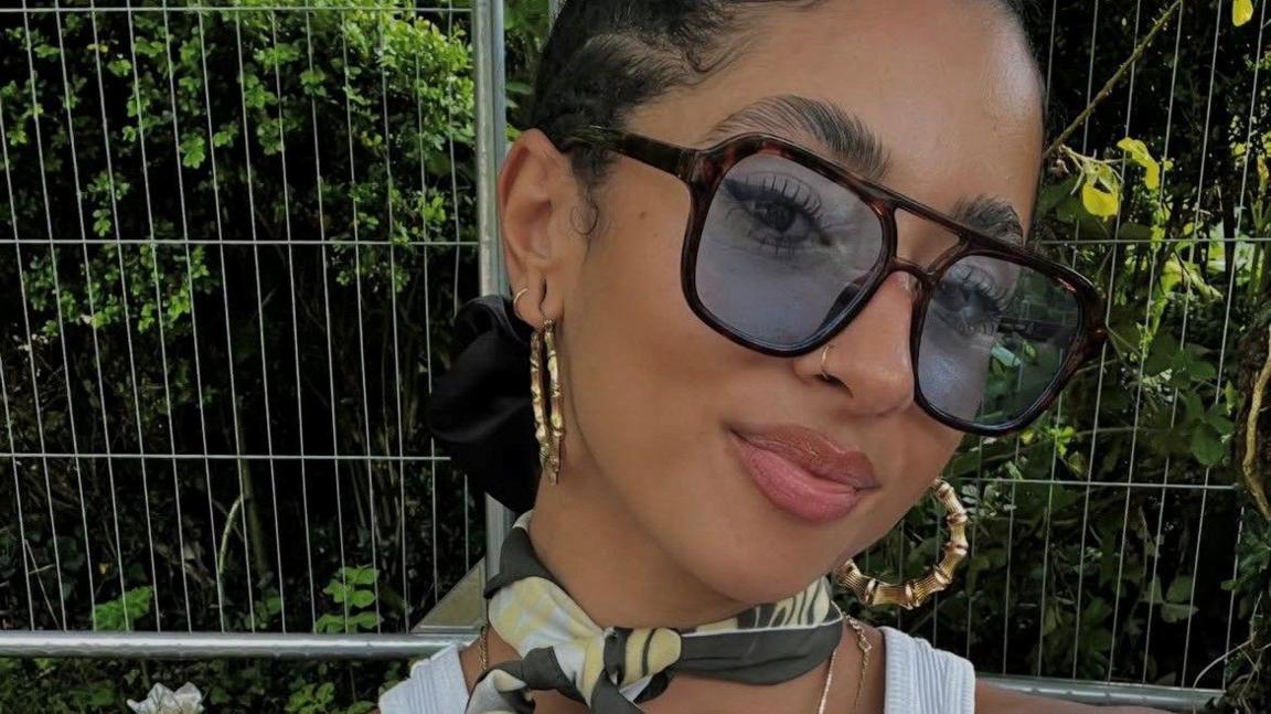 A woman is wearing large sunglasses and earrings, and is looking at the camera.