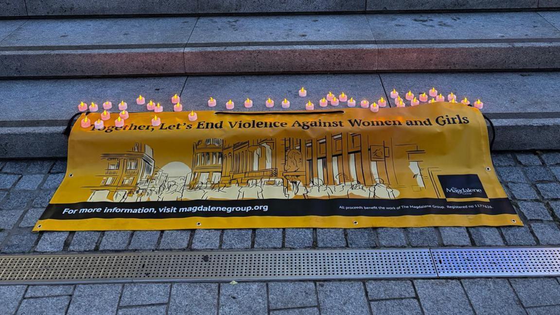 A yellow banner placed on concrete steps. It reads "Together, Let's End Violence Against Women and Girls". There are multiple candles on the upper step.