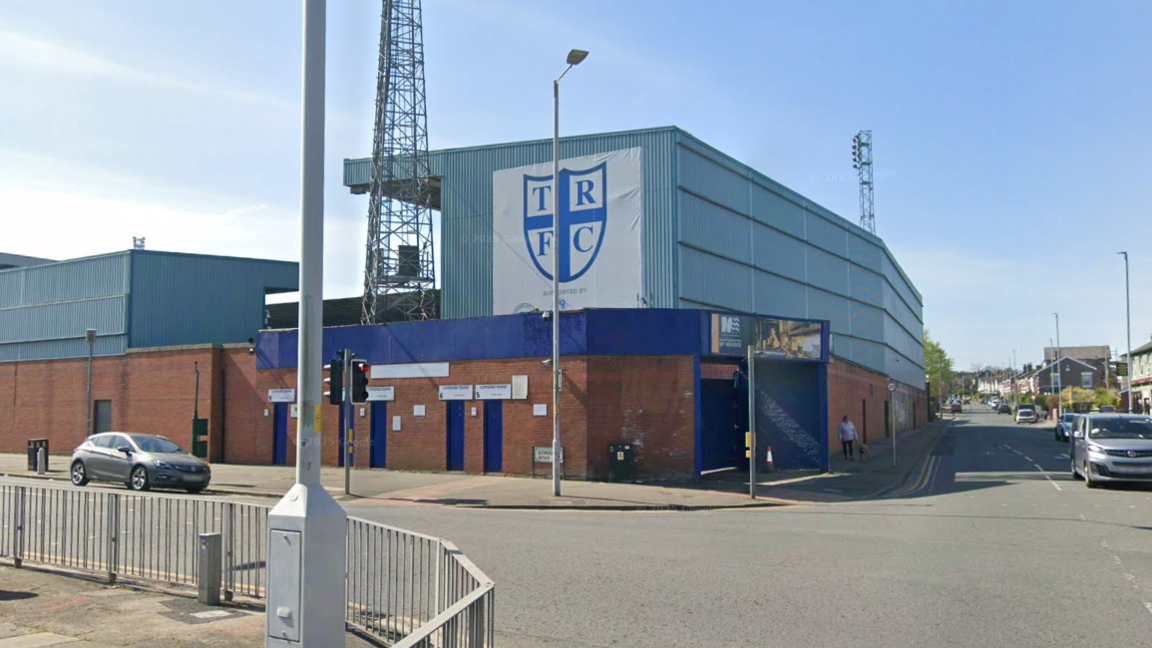 Google Streetview image of Tranmere Rovers' Prenton Park stadium. Taken from outside the ground, two large stands are visible. One of them includes a large blue-and-white shield with the initials TRFC printed on it.