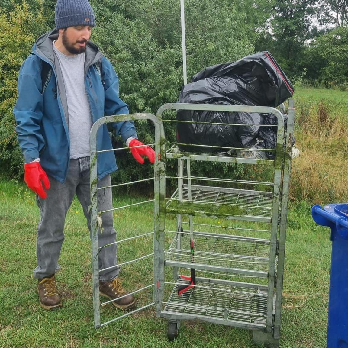 A man stands in a meadow looking at a large metal trolley rack with black bags of rubbish on top. He has a beard and wears a blue jacket, light top, blue hat, grey jeans and red gloves. wearing grey jeans, red gloves and brown shoes.