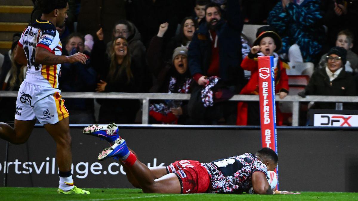 Leigh centre Tesi Niu diving over to score a try with supporters jumping up in celebration in the background. Catalans Dragons' Phoenix Laulu-Togaga'e is in the foreground