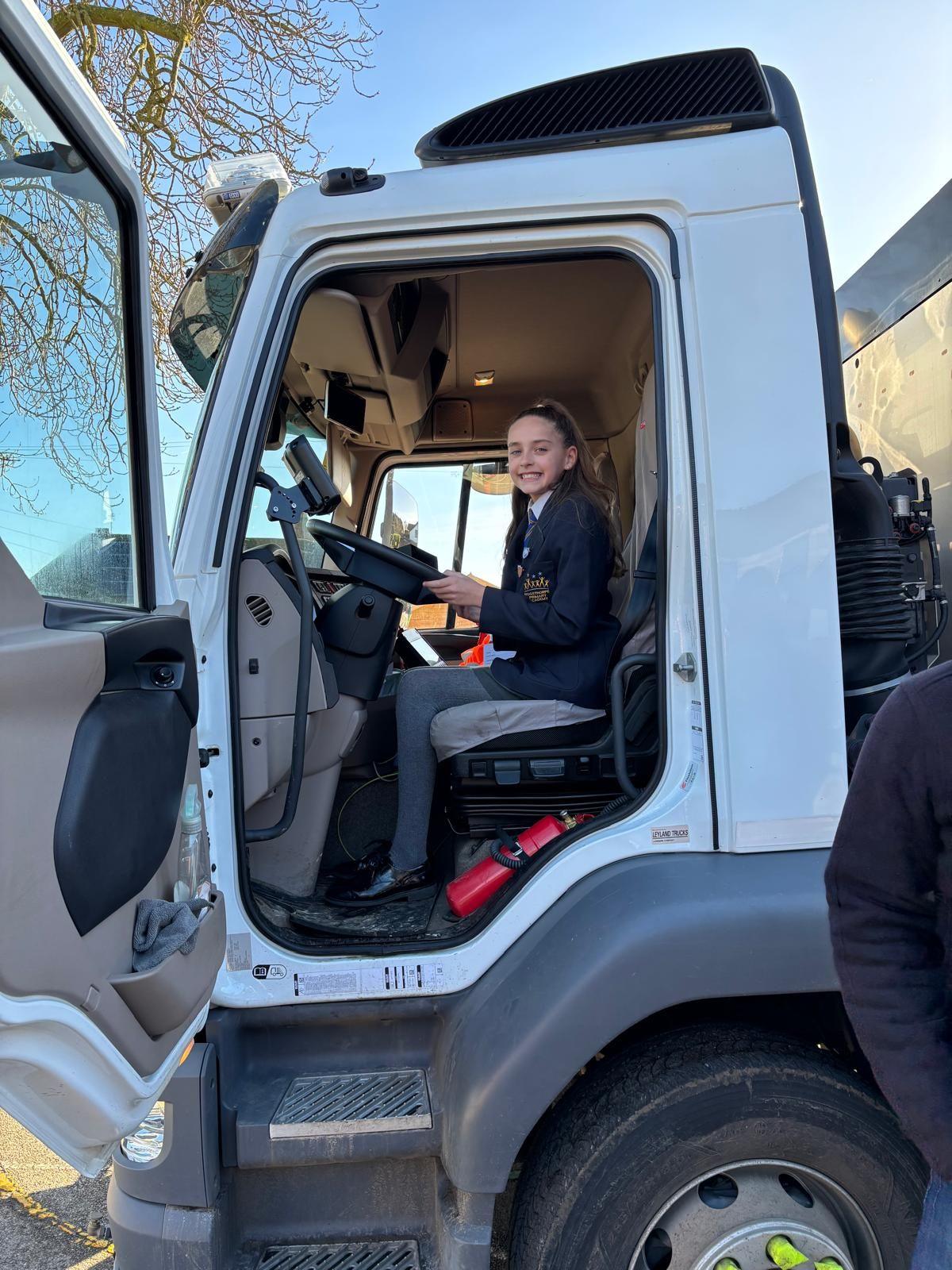 One of the girls, Brooke, sitting inside the cab of a road sweeper. The front door is open. She is holding the steering wheel and smiling at the camera.