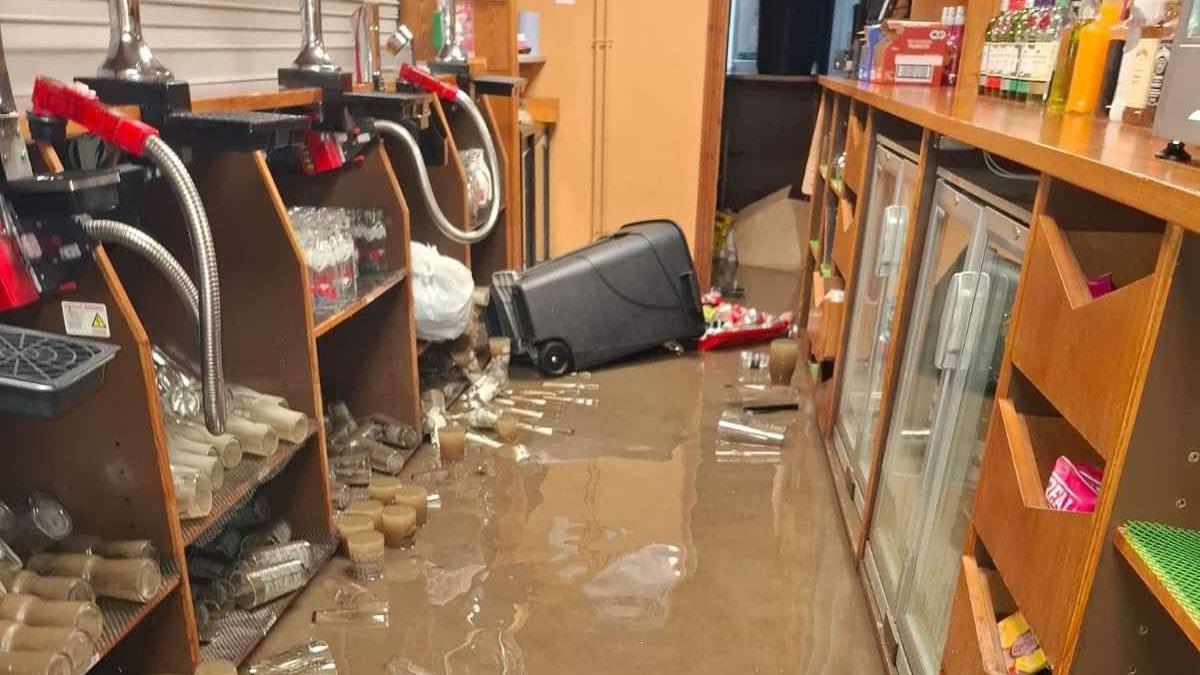 A flooded bar is seen with a few centimetres of water visible. A bin is on its side with  several items scattered floating in the water.