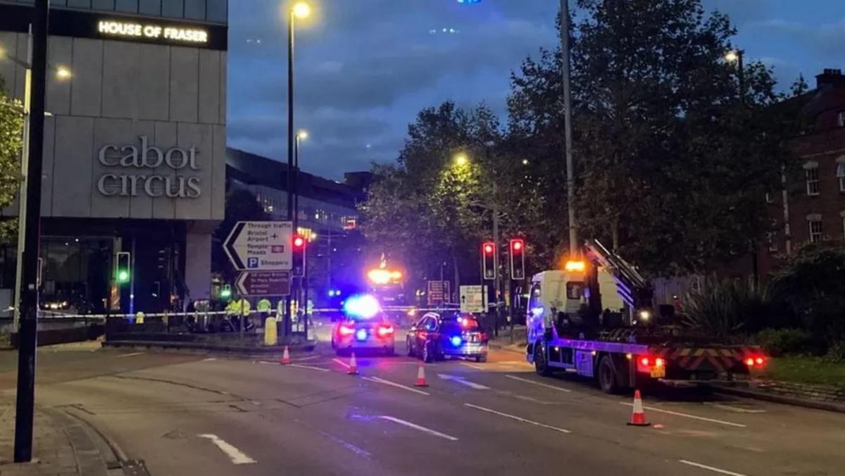 Cabot Circus shopping centre at night. There are several police cars parked next to it with their flashing lights on, and a police cordon is in place at the scene of the crash.