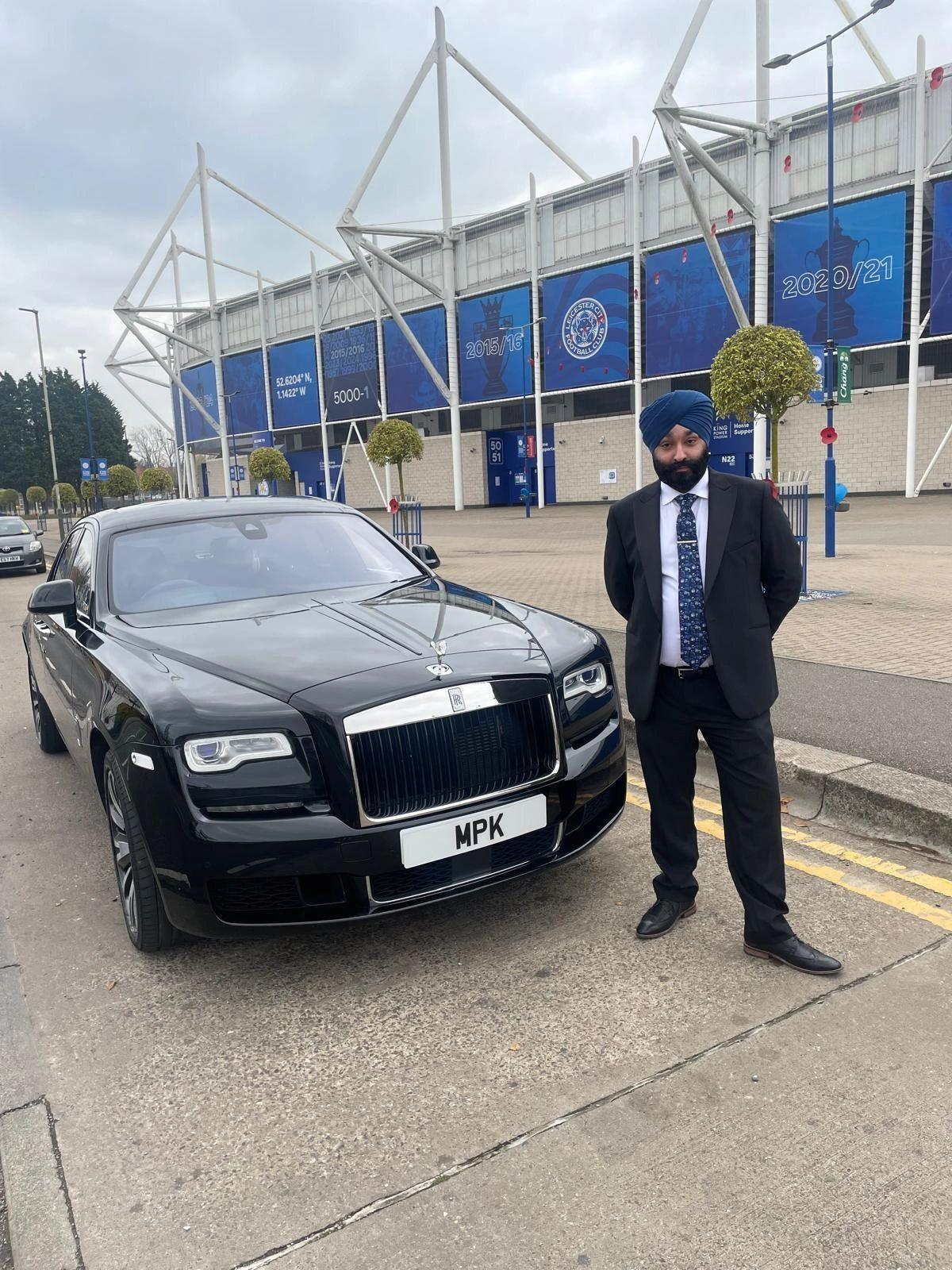 Tony Singh is wearing a business suit and a turban. He is standing in front of Leicester City Football Club. Behind him is a luxury car with the number plate MPK. 