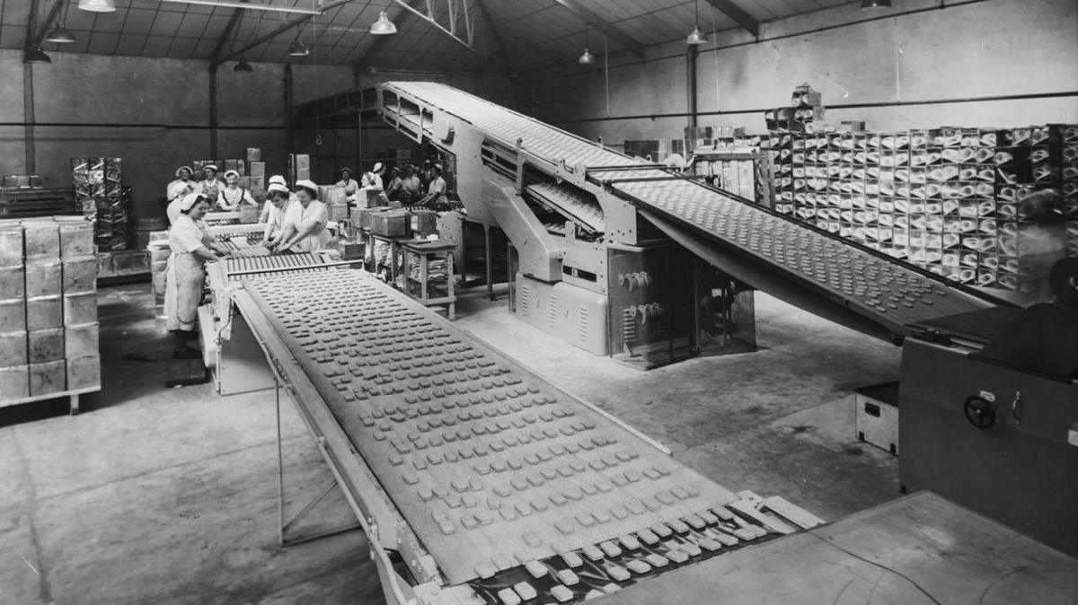 A black and white photo of workers packaging the biscuits at the Hill bakery in Ashton-under-Lyne. Dozens of people are working on the production lines.