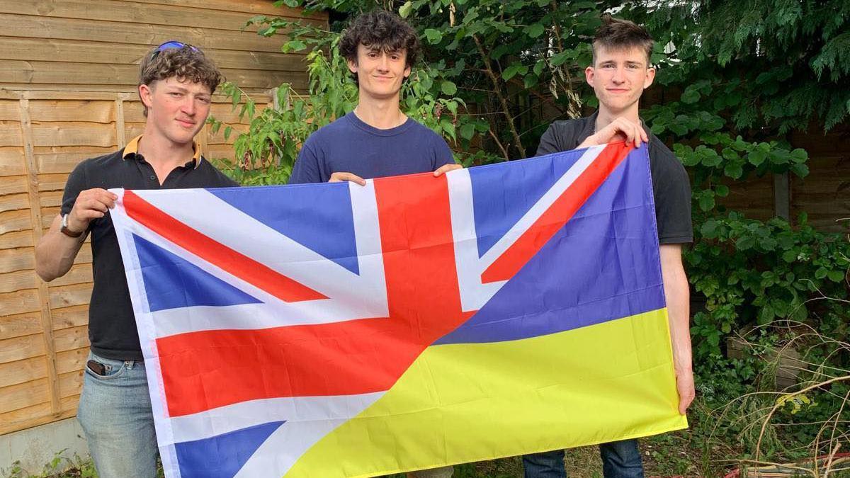 Kyle Spring (L), James McCarthy-Hill, and Adam Mitchell (R) smiling and holding up a large flag which is split diagonally across the middle. The top half is the Union flag, and the bottom half is the blue and yellow Ukrainian flag. They are standing in a garden in front of a wooden fence and some greenery.