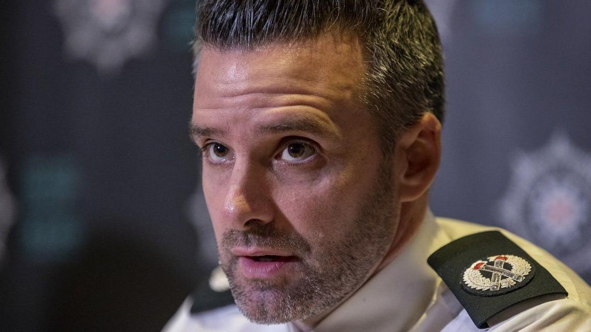 A police officer in close up wearing his uniform shirt, with epaulettes on the shoulders. He has short, thick salt and pepper hair and stubble. 