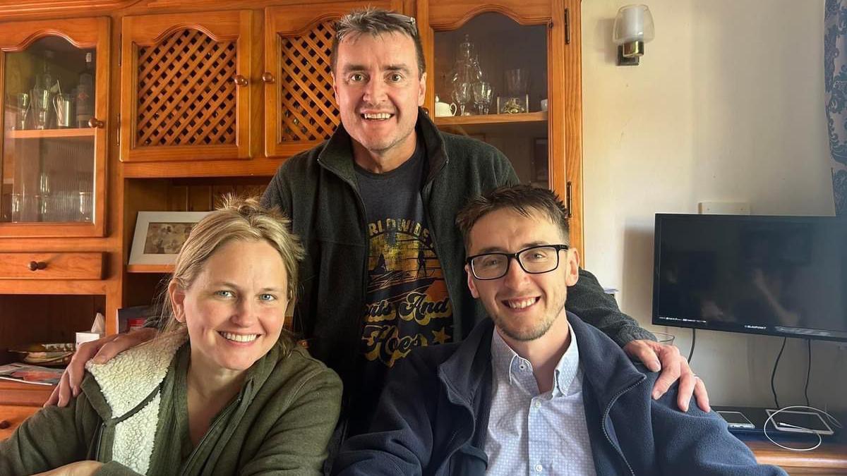 Three people round a table smiling. There is a woman on the left hand side wearing a gree hoodie with blonde hair. there is a man standing in the middle who is wearing a graphic tshirt and smiling with a pair of glasses on his head. There is a man on the right hand side wearing a light blue spotted shirt and a light blue fleece jacket