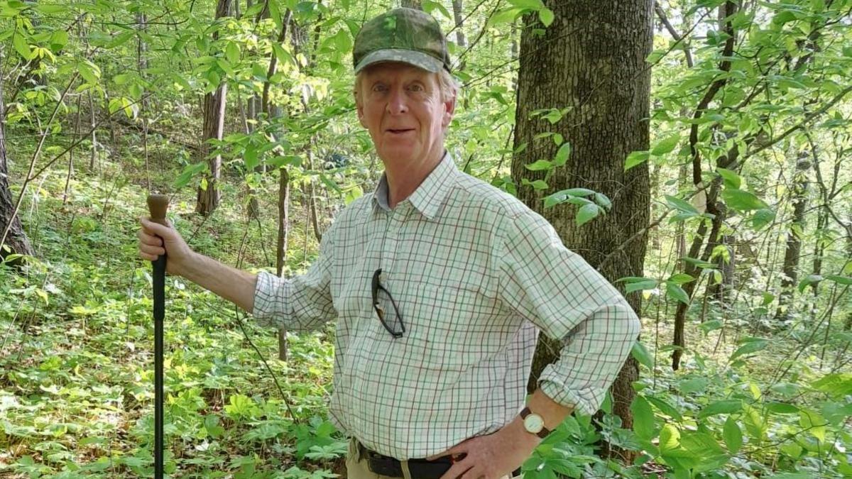 Robert Benson, a middle-aged man wearing a checked shirt and a cap, is standing with trees and foliage behind him 