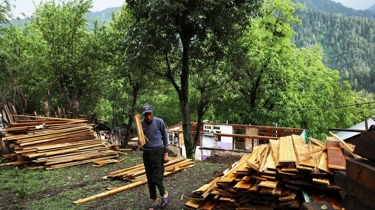 A man in a cap and blue jumper carries a plank of wood, others laid out either side of him and a destroyed house behind him. There is a lush valley in the background.