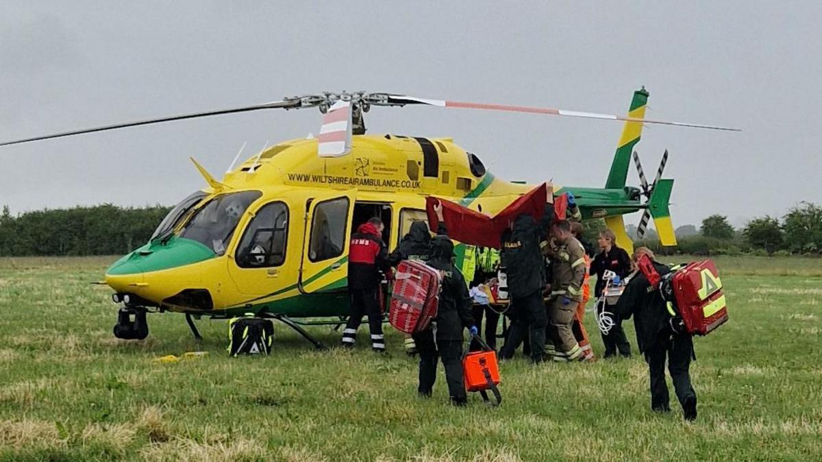 A Wiltshire Air Ambulance has landed in a field. Paramedics and fire crews are seen gathered around the helicopter.