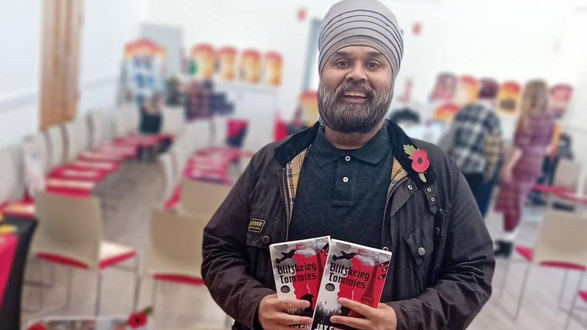 Jax Singh Taak is looking at the camera and smiling. He is holding two copies of his book, which shows a black blane over a grey-and-white map of Europe featuring the Eiffel Tower and the title "Blitzkreig Tommies". He is wearing a dark Barbour-style jacket, a dark green polo shirt, a light grey turban and a red poppy. In the background are chairs, tables and the backs of two women.