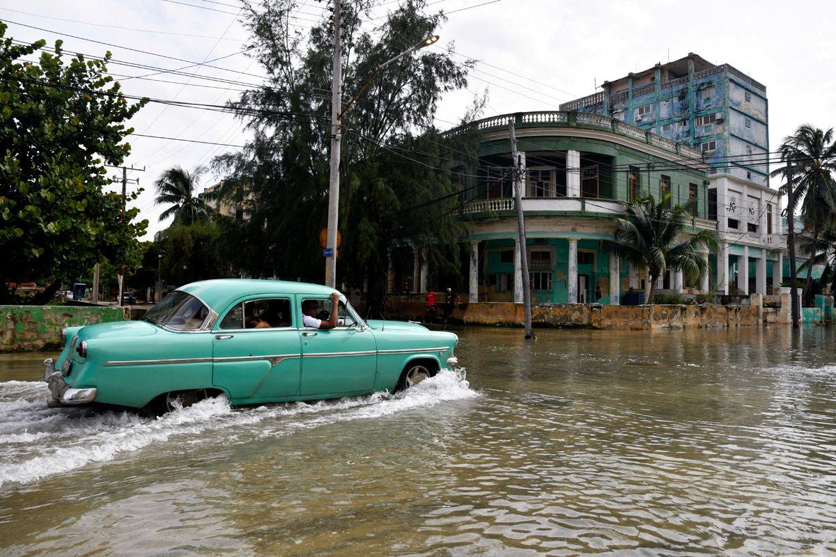 A vintage green car crosses a flooded street with buildings and palm trees in the background in Havana, Cuba, October 9, 2024