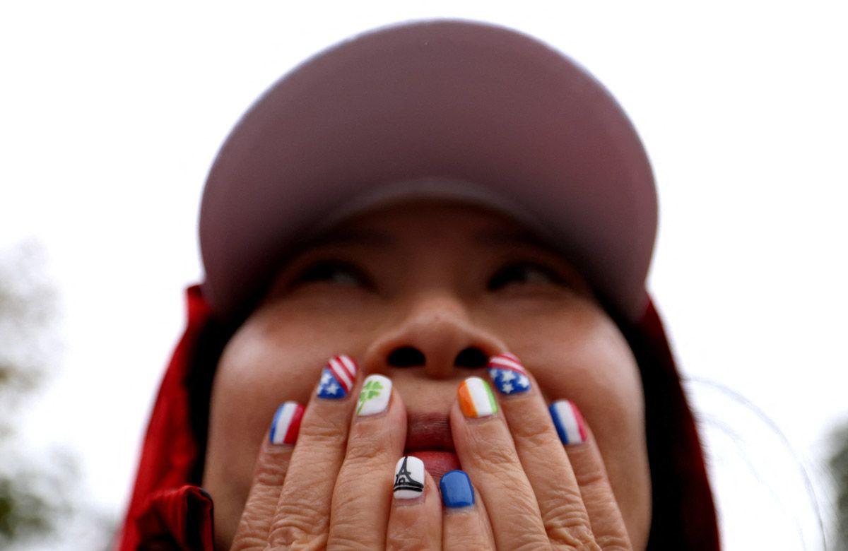  A woman wearing a cap covers her mouth with her hands. Her nails are painted with  flags and symbols