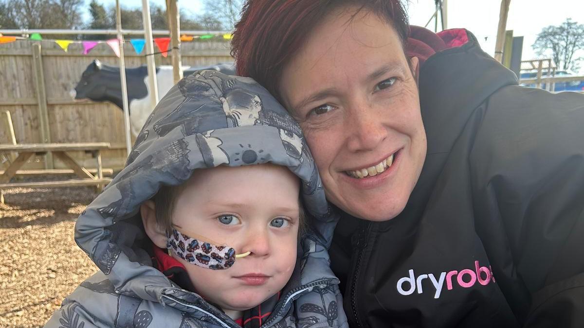 A selfie showing Connor wearing a grey patterned raincoat with a feeding tube in his right nostril, secured with a large plaster with lorries on it. He is sitting beside his mum Lucy, who has short dark red hair and is smiling at the camera. They appear to be sitting in a play park with bark chippings on the ground and colourful bunting hanging behind them.