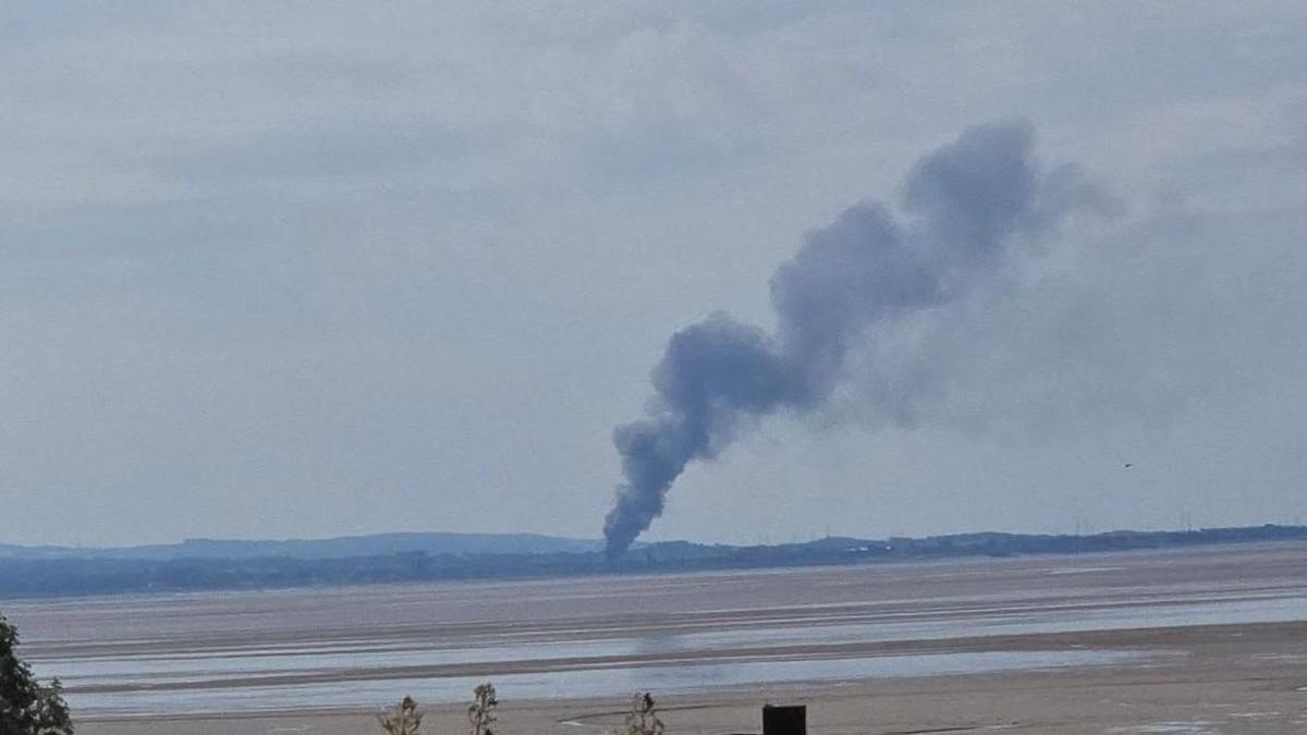A large plume of smoke can be seen billowing above the sands of Morecambe Bay with hills in the background.