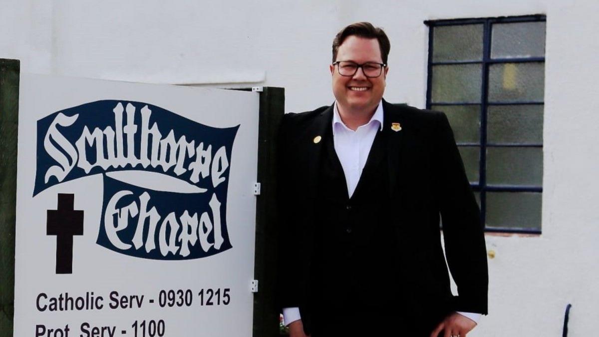 Ian Brown is wearing a black suit and white shirt. He is standing in front of a sign that says "Sculthorpe Chapel".